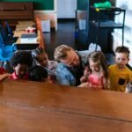 A warm classroom scene showing a teacher interacting with diverse preschool children around a piano.